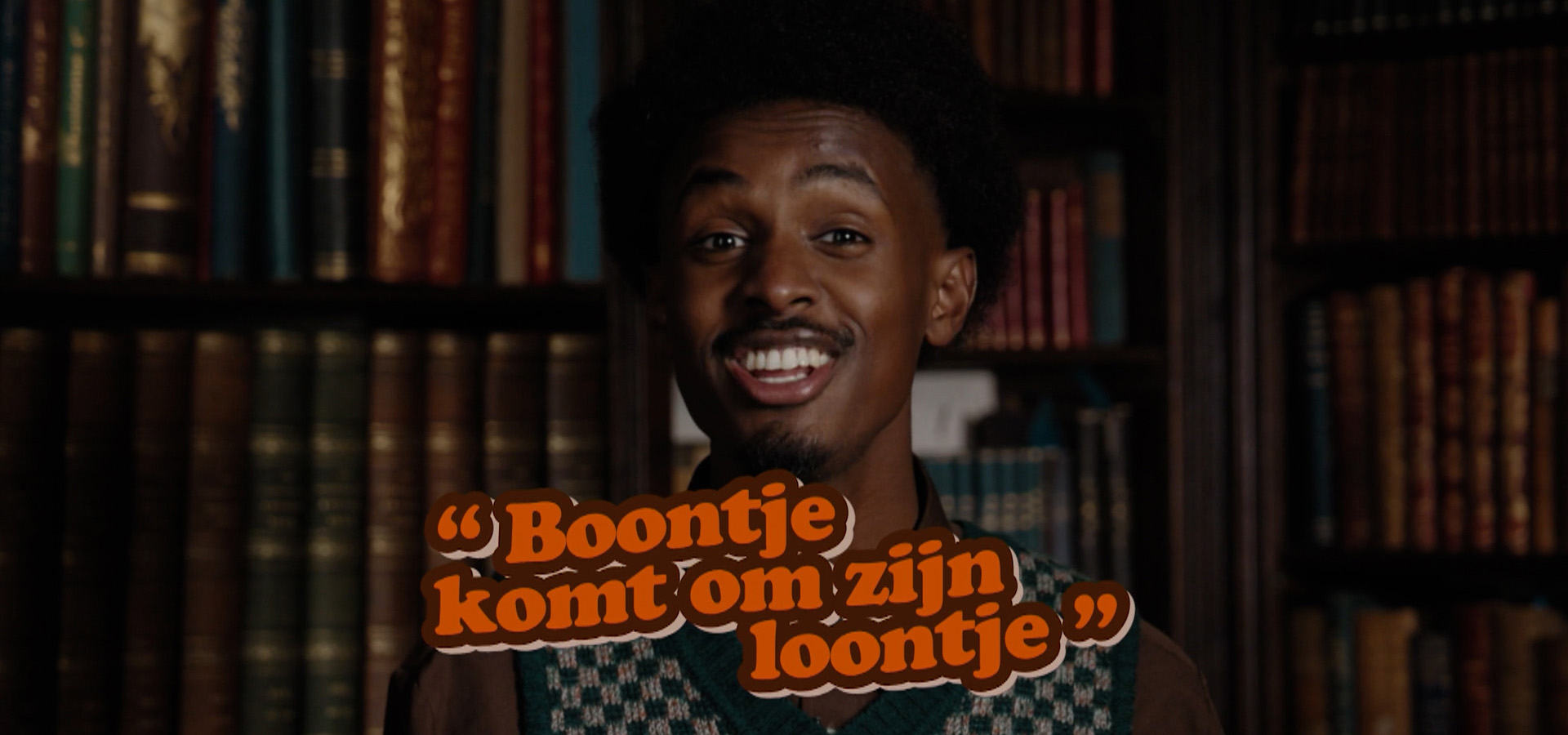 Foto van presentator Anas voor boekenkast met tekst 'boontje komt om zijn loontje'.